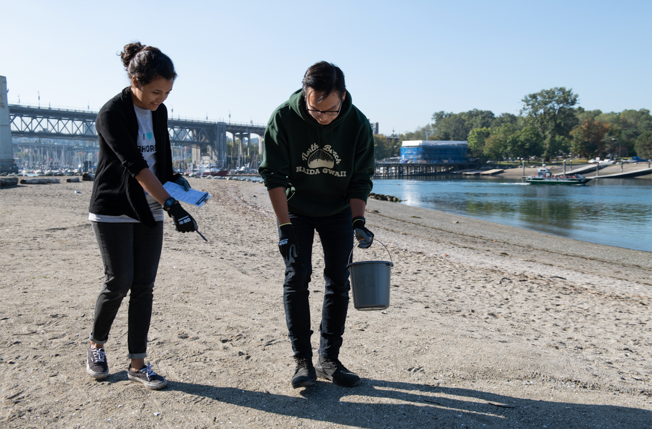 A man and woman walk on the beach collecting data on plastic using a notepad and a small bucket. - click to enlarge A man and woman walk on the beach collecting data on plastic using a notepad and a small bucket.