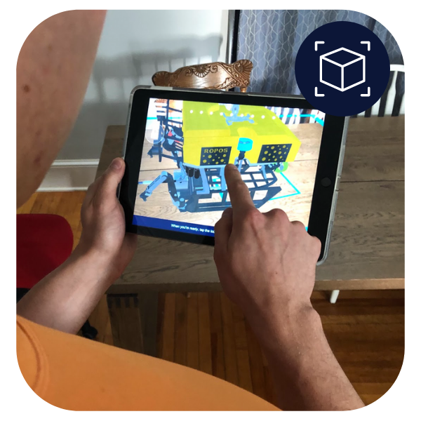 Person looking at the Fly ROPOS experience on a tablet. The augmented reality icon used on the Ocean School website is in the top right corner of the image. This icon shows a cube. - click to enlarge Person looking at the Fly ROPOS experience on a tablet. The augmented reality icon used on the Ocean School website is in the top right corner of the image. This icon shows a cube.